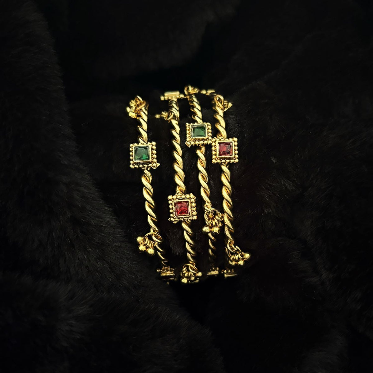 Two pairs of Jhanak twisted brass kada bangles stacked, showing red and green glass settings and ghungroo bells