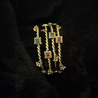 Two pairs of Jhanak twisted brass kada bangles stacked, showing red and green glass settings and ghungroo bells