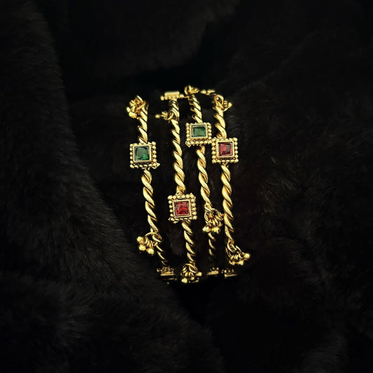 Two pairs of Jhanak twisted brass kada bangles stacked, showing red and green glass settings and ghungroo bells